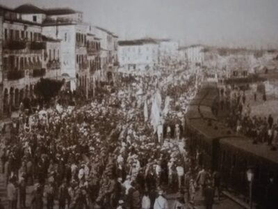 Η γιορτή των Ελευθερίων στην Πάτρα, τη δεκαετία του 1910. Πλήθος κόσμου έχει συρρεύσει στο λιμάνι ενώ στη μέση υπάρχει αμαξοστοιχία για τη μεταφορά των επιστήμων και επισκεπτών/mixanitouxronou.gr